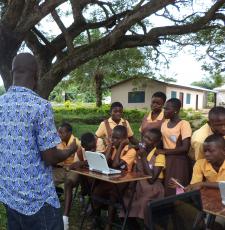 A librarian teaching a group of school children to learn computer skills. The class is taking place outside, under a tree.